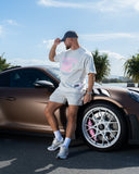 Man leaning against a brown sports car with palm trees and blue sky in the background