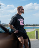 [men] Man wearing a washed black EVDNCE 02 shirt with pink text by a Porsche GT3 RS by a waterfront.