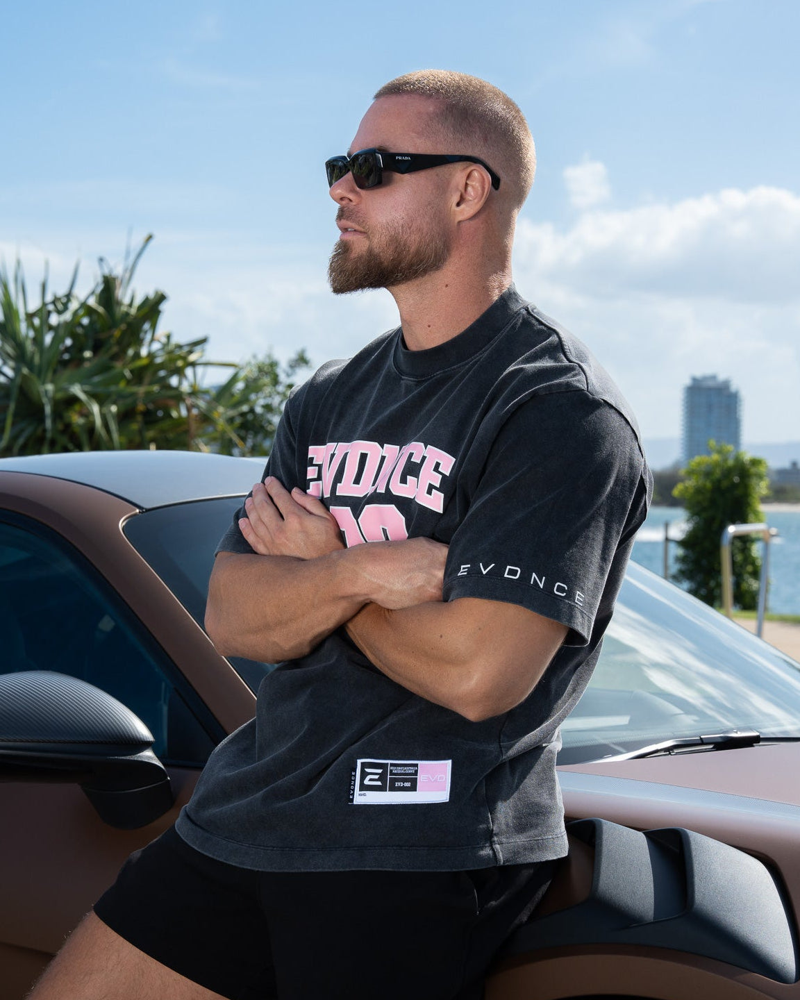 Man wearing a washed black EVDNCE 02 Tee with visible branding, standing next to a car with a scenic background.